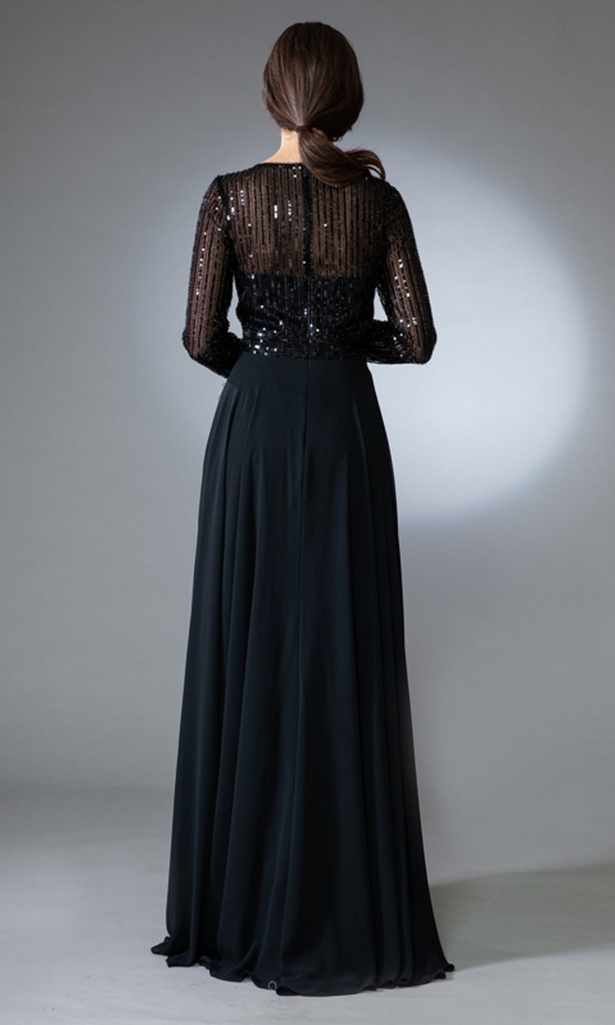 A woman stands with her back to the camera in Long-Sleeve Long Modest Prom Dress 7036, featuring a sequined semi-sheer top and flowing skirt—a stunning evening gown against a plain, light background.